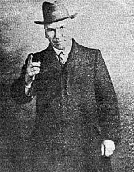 black and white photo of a man wearing a hat and coat pointing his index finger towards the camera.