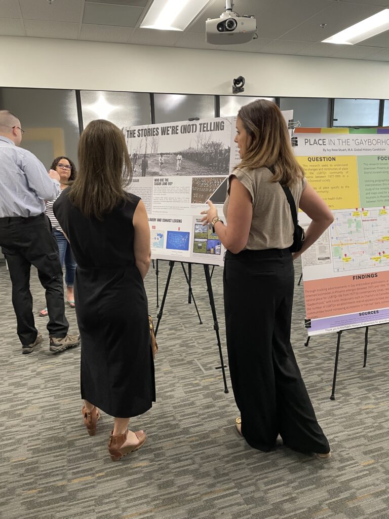 Two women stand in front of a poster facing it and talking.